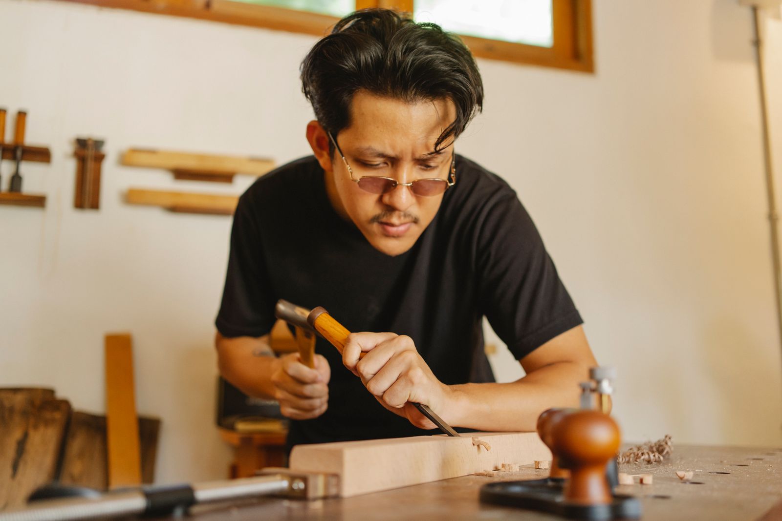 Concentrated Asian man working with chisel and hammer while cutting notch in hardwood plank
